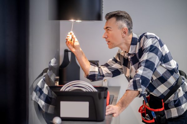 Renovation. Profile of concentrated middle aged man in plaid shirt touching screwdriver to lamp on kitchen range hood in modern kitchen at home
